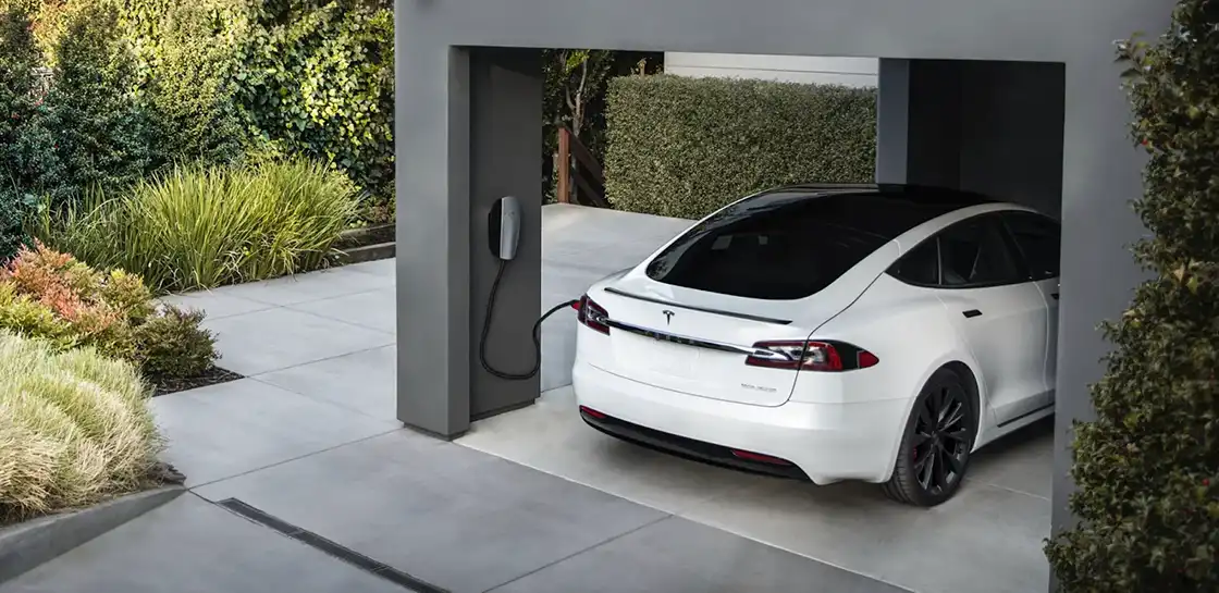 A white tesla electric vehicle seen parked in an open garage connected to a charger